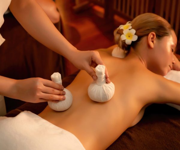 Hot herbal ball spa massage body treatment, masseur gently compresses herb bag on woman body. Tranquil and serenity of aromatherapy recreation in warm lighting of candles at spa salon. Quiescent