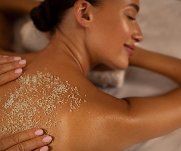 Serene woman deeply relaxing as spa professional applying an exfoliating treatment to her back, promoting wellness and skin health, top view